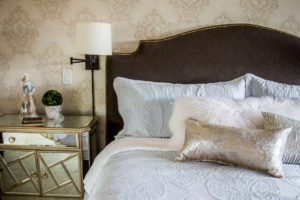 section of master retreat showcasing subdued wallpaper, custom headboard, wall sconce and mirrored chest